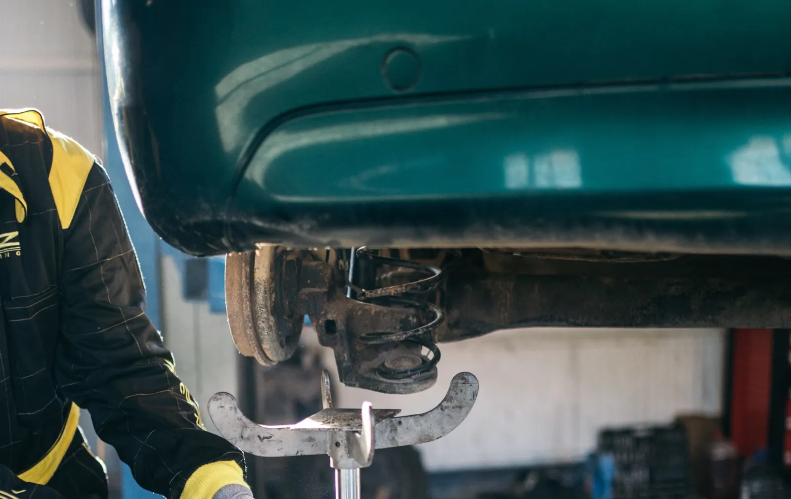 A mechanic beside a raised car in a repair shop, with a worn, rusty rear axle and coil spring above a jack stand