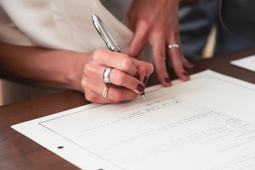 A hand holding a silver pen signs a printed form on a wooden desk