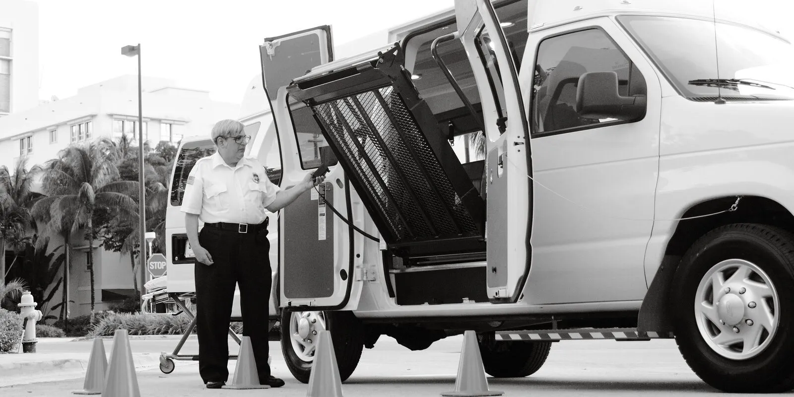 A driver in a uniform shirt holds the hand control as a wheelchair lift folds into the side door of a white high-roof van