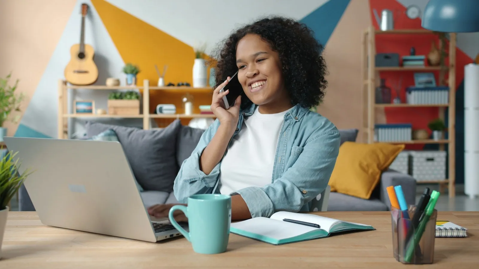 A smiling woman talks on her phone at a desk in her living room, with a laptop, an open notebook, and a mug in front of her