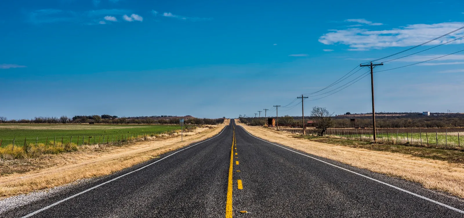 A two-lane country highway running straight through flat Texas farmland under a blue sky, with power lines along one side