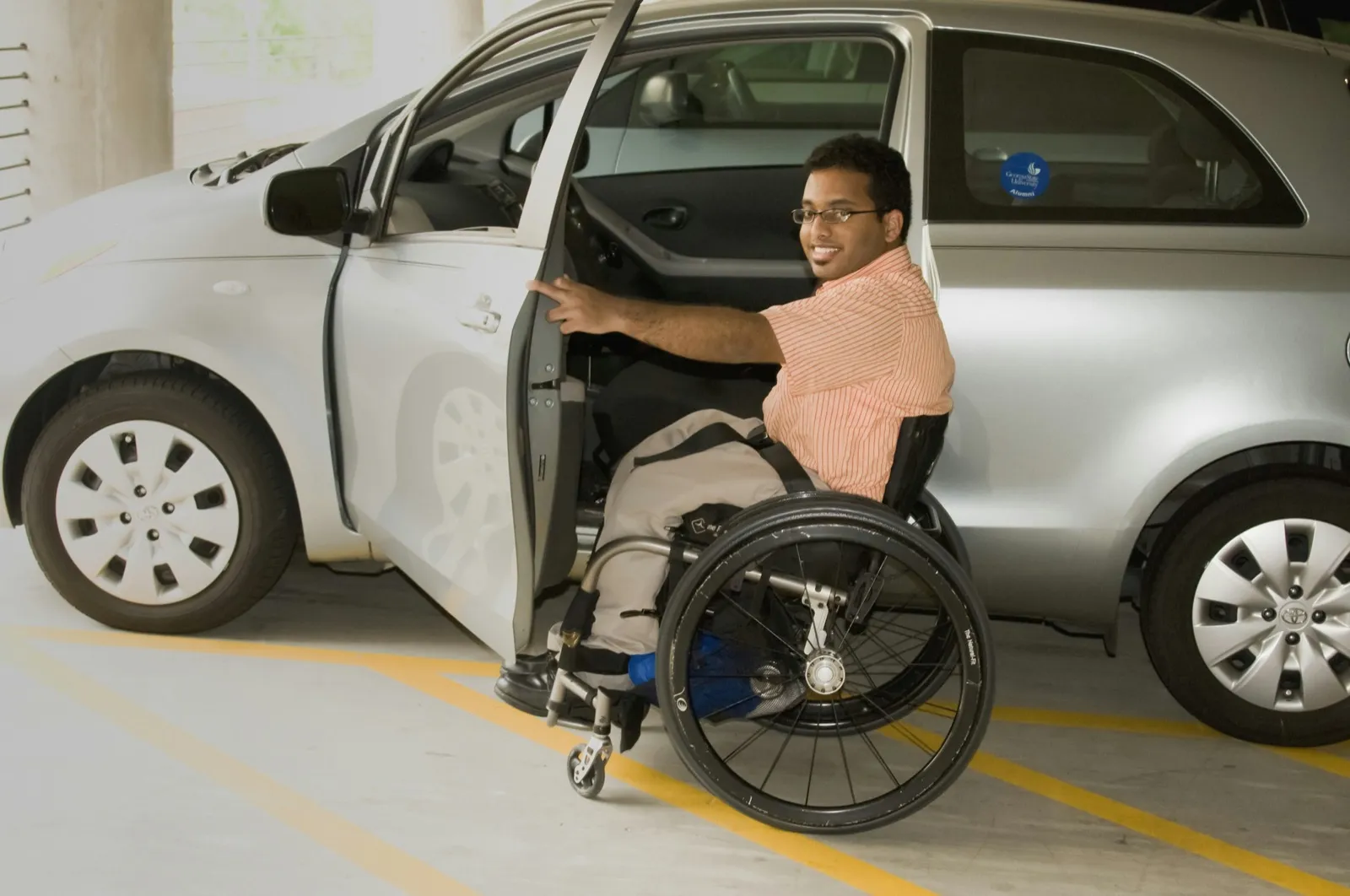 A man in a manual wheelchair holds the open door of a small silver car in a parking garage, lined up to move from his chair to the seat