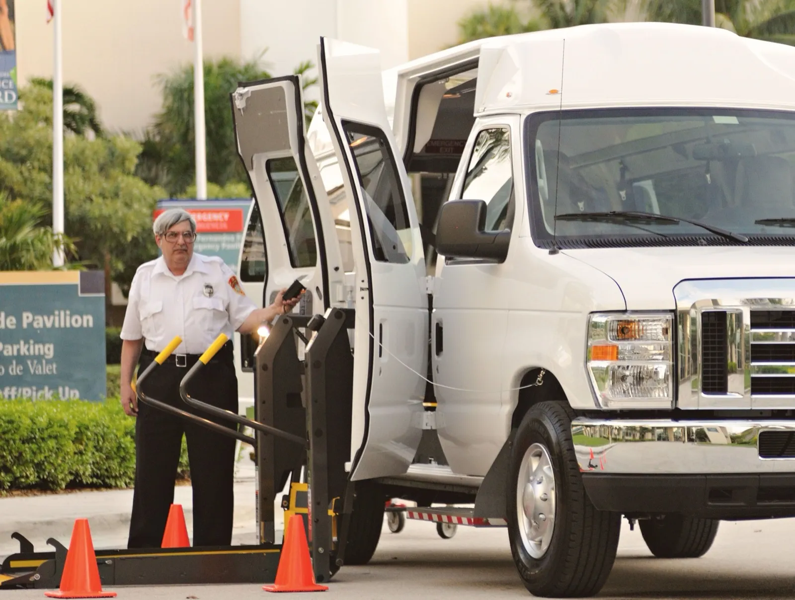 A driver in a white uniform shirt stands at the open side lift of a white wheelchair van parked at a hospital entrance