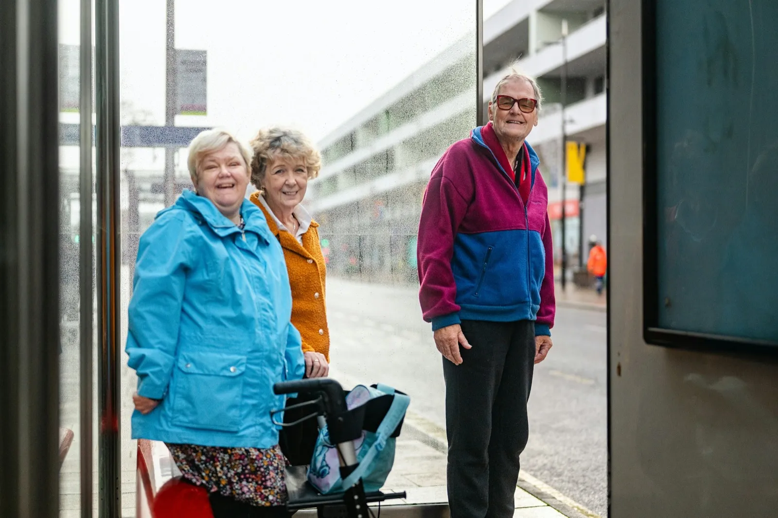 Three smiling older adults wait together at a glass bus shelter on a city street, one resting her hands on a rolling walker