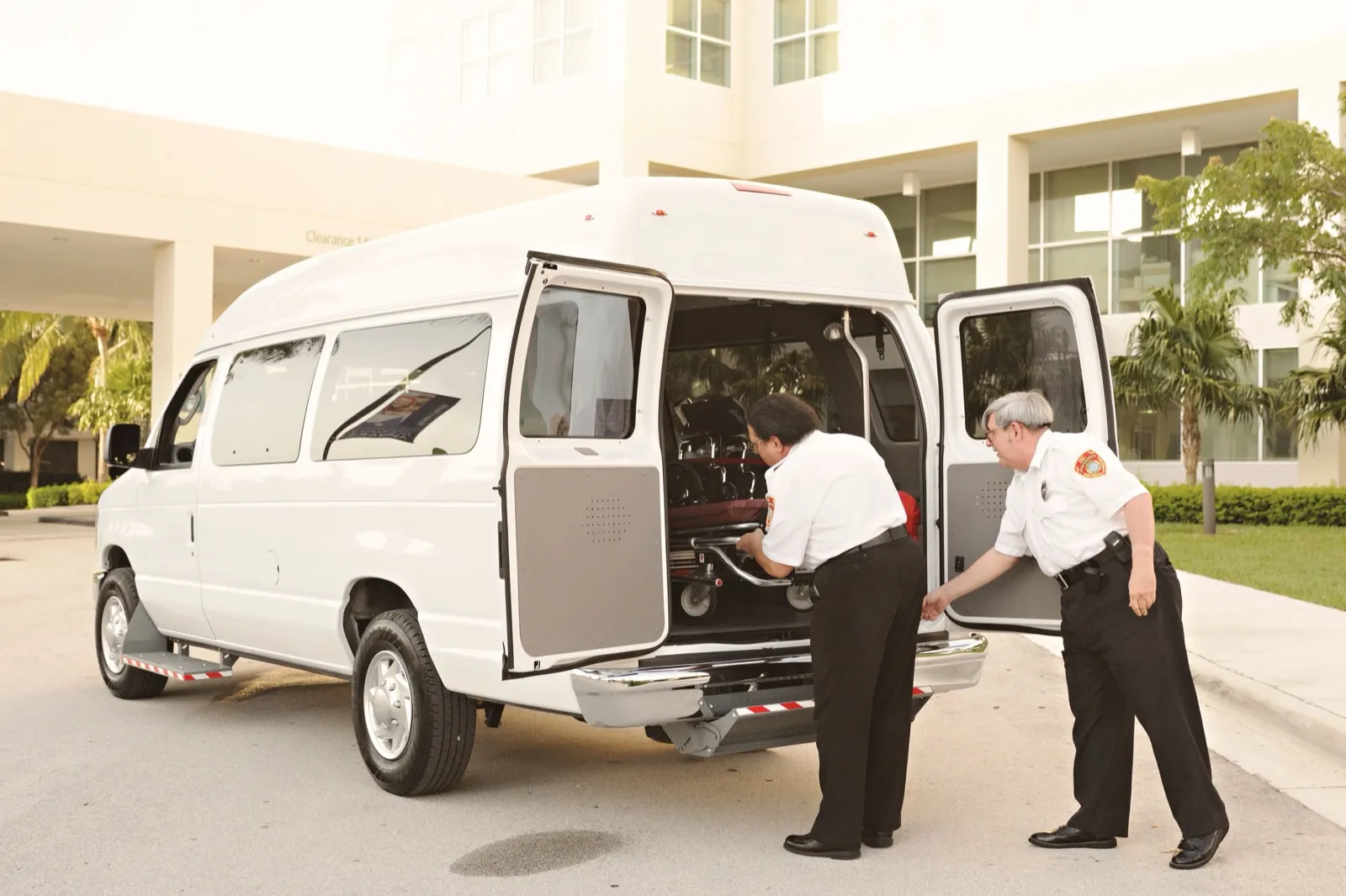 Two crew members in white uniform shirts load a wheeled stretcher through the open rear doors of a white high-roof van outside a medical building