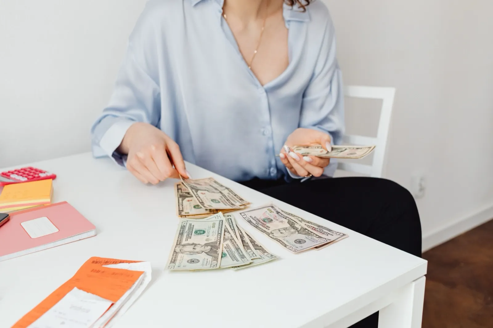 A person at a white table counts a few U.S. dollar bills, with a calculator, a notebook, and receipts beside them
