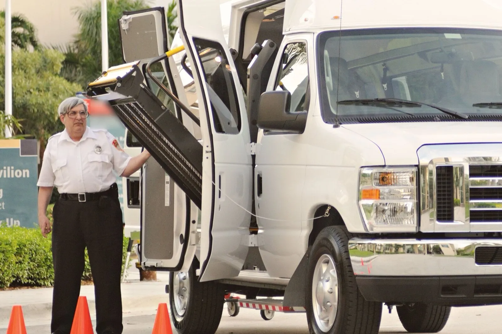 A man in a white uniform shirt stands beside the open side doors and raised wheelchair lift of a white high-roof van