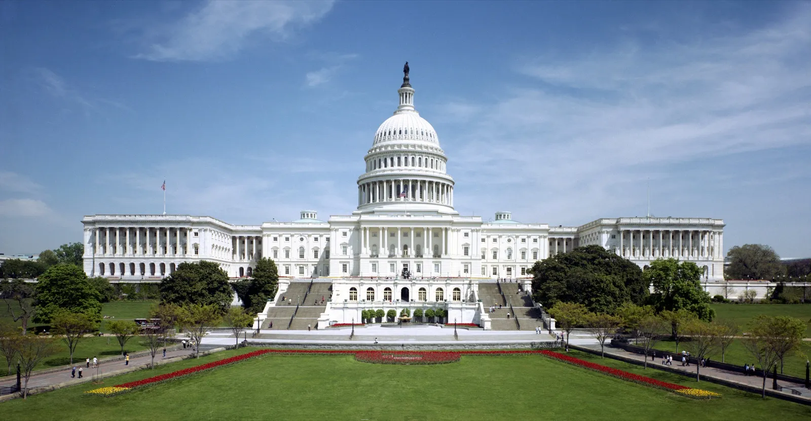 The west front of the United States Capitol in Washington, D.C., with its white dome, wide steps, and green lawn under a blue sky