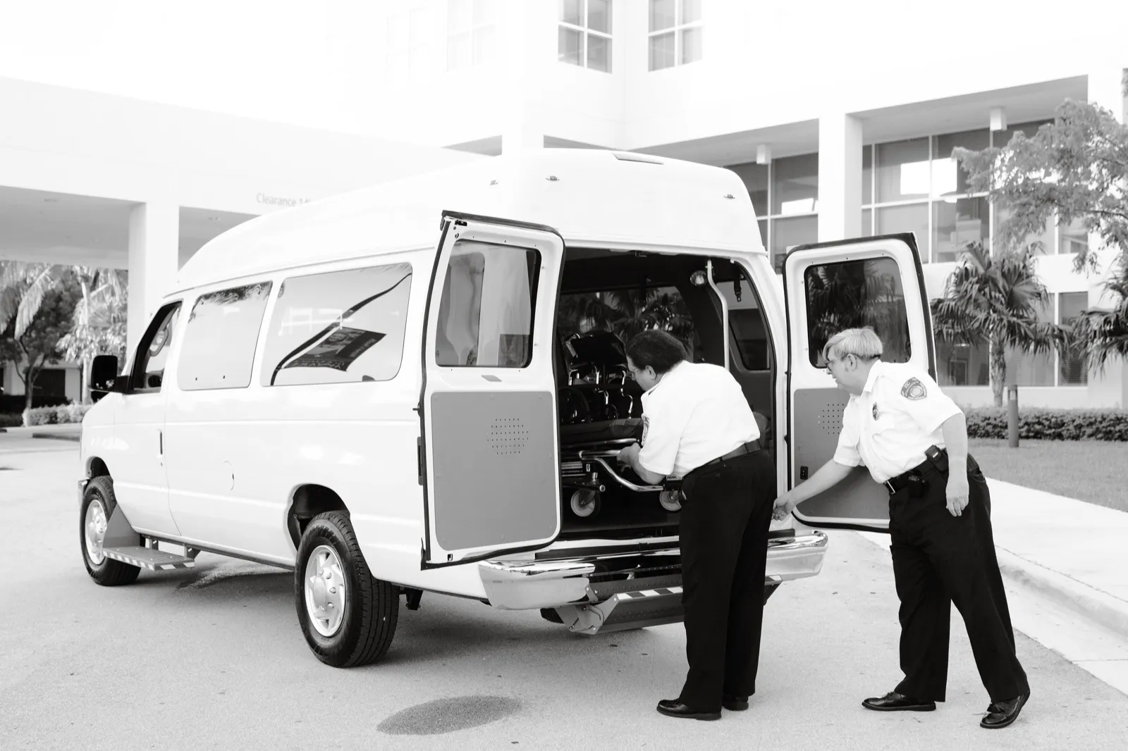 Two crew members in uniform shirts load a wheeled stretcher through the rear doors of a white high-roof van outside a hospital, in black and white