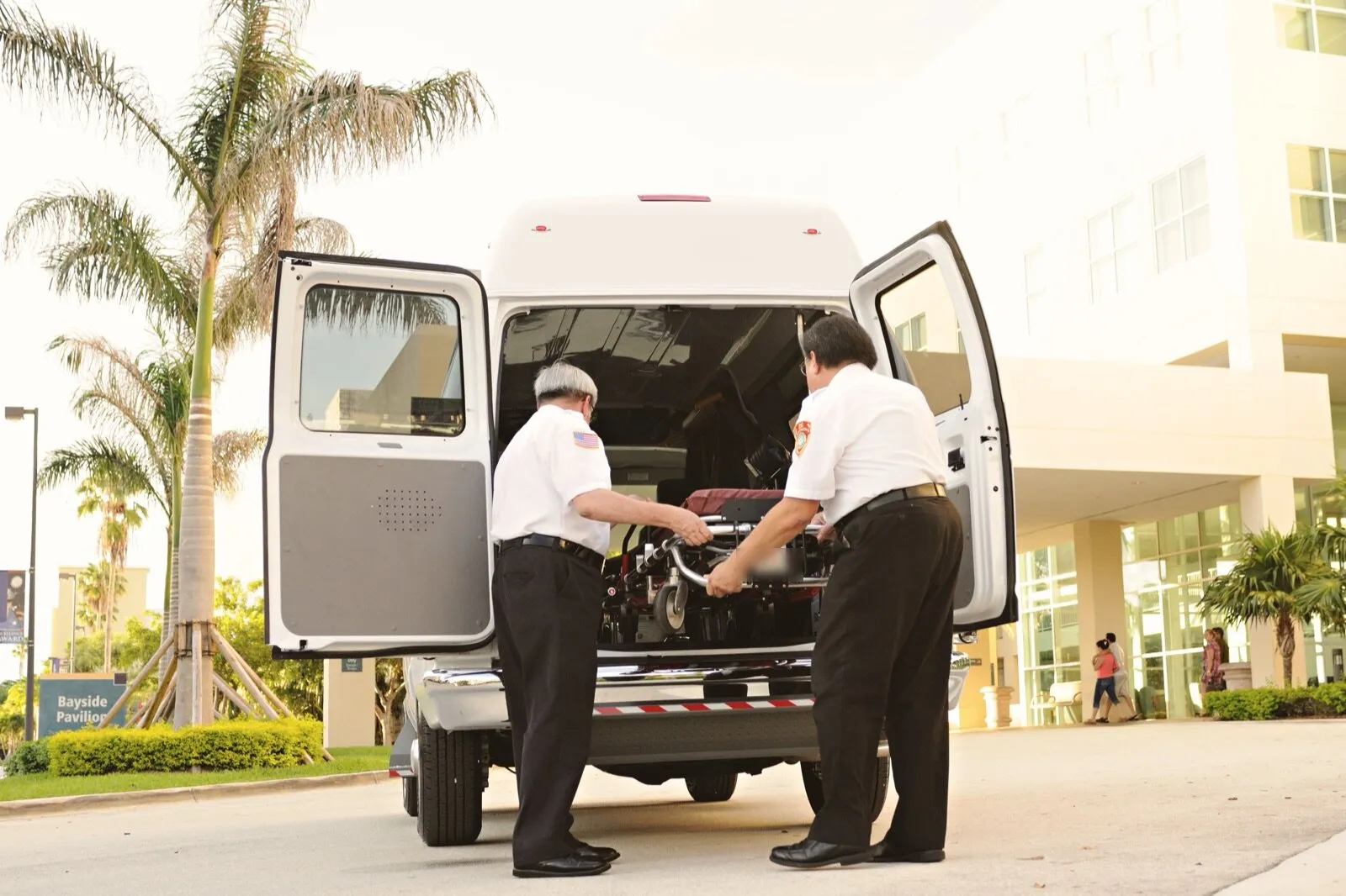 Two crew members in white uniform shirts load a wheeled stretcher through the open rear doors of a white high-roof van outside a hospital