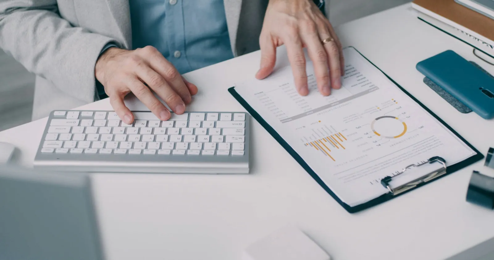 Hands typing on a white keyboard beside a clipboard holding a printed report on an office desk