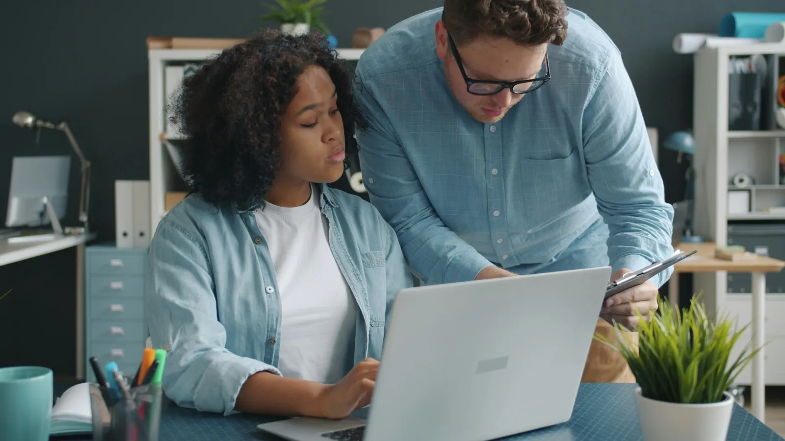 An office worker types on a laptop while a coworker leans in beside her holding a clipboard