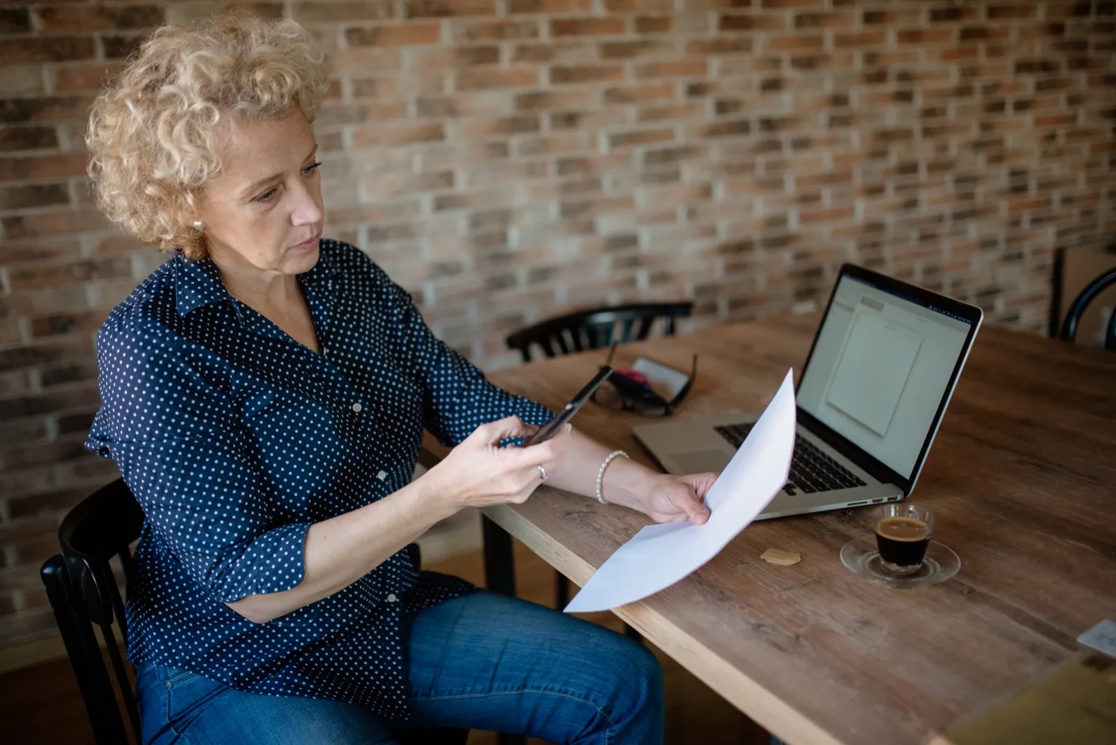A woman at a wooden table photographs a paper document with her phone, next to an open laptop and a cup of espresso