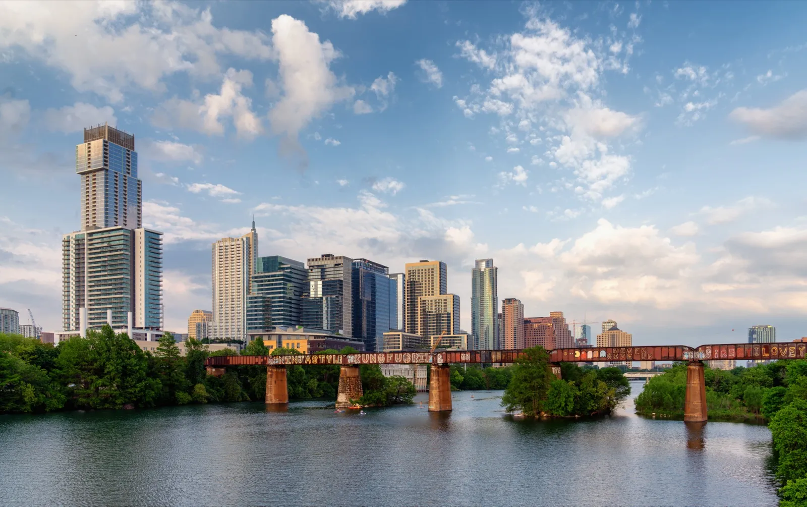 The downtown Austin skyline on a sunny afternoon, seen across Lady Bird Lake with an old railroad bridge and green trees in front