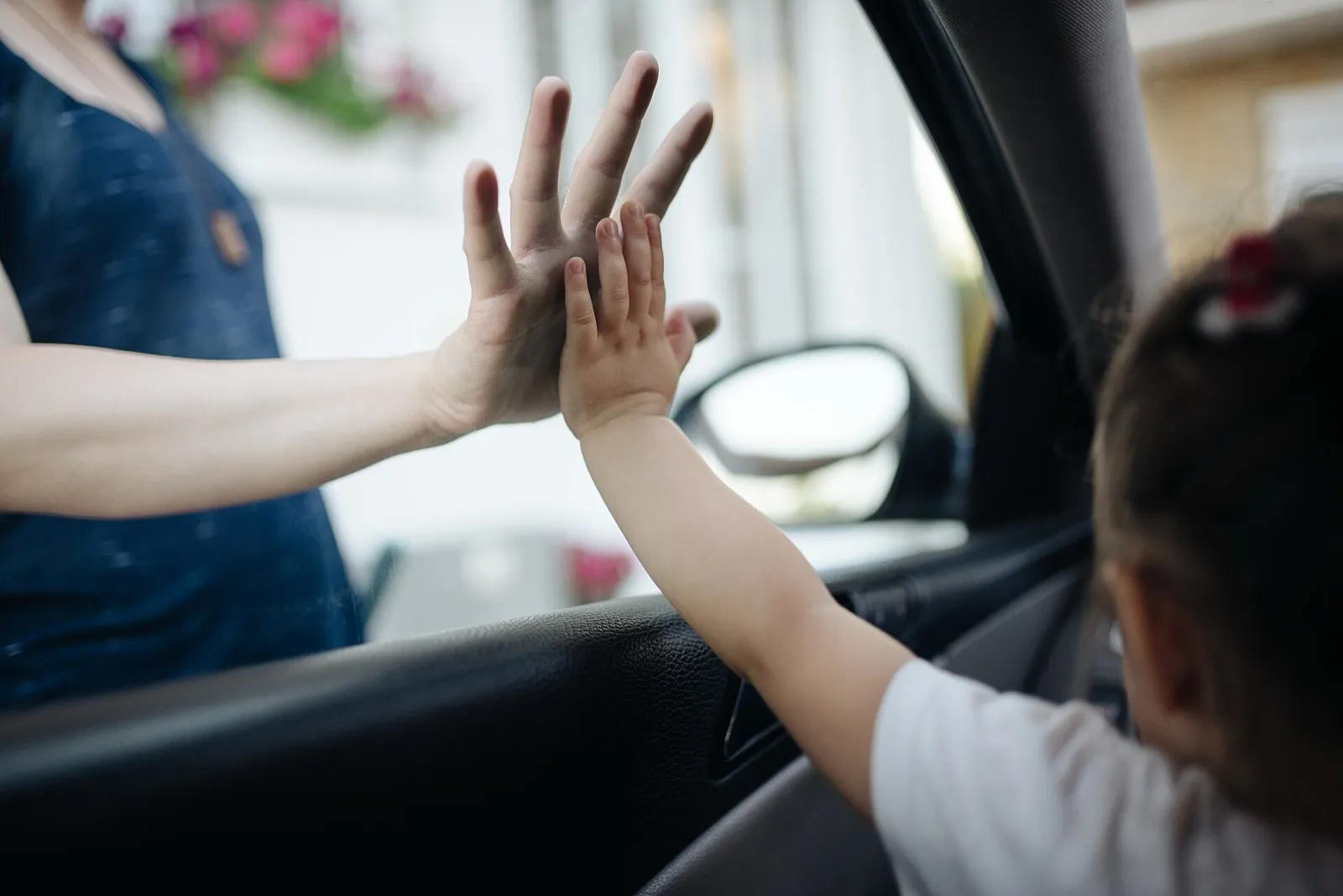A child inside a car, seen from behind, presses a hand against an adult's hand at the car window