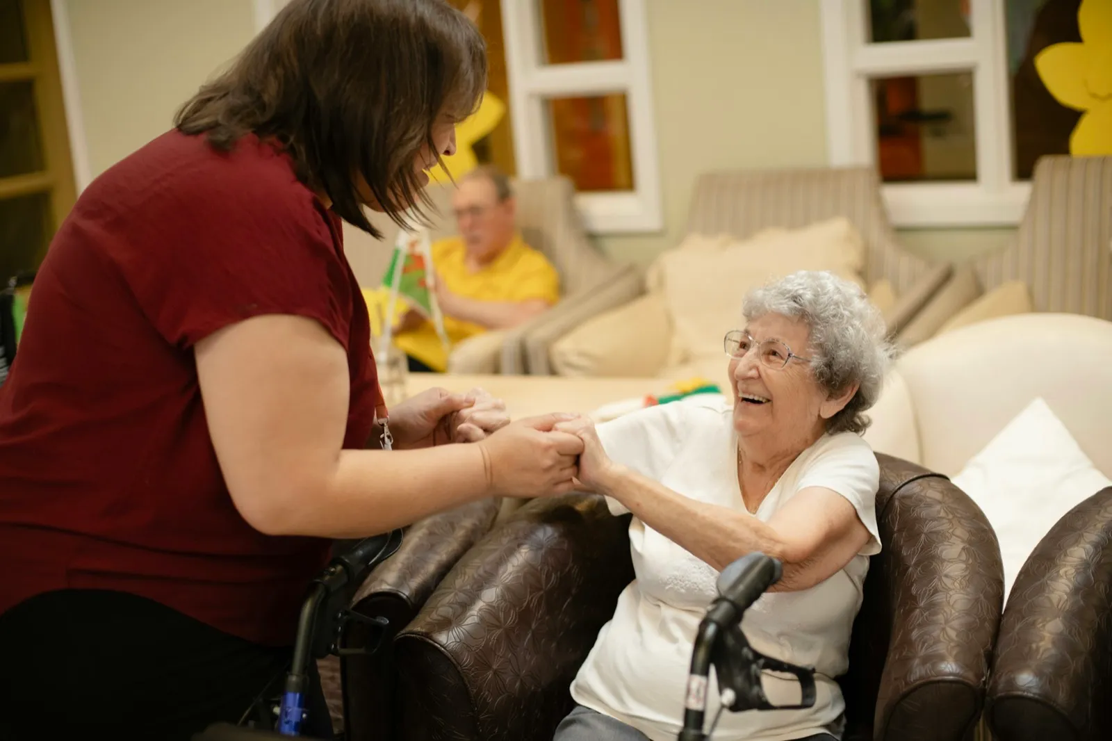 A caregiver holds the hands of a smiling older woman seated in an armchair with a walker in front of her, in a care home lounge
