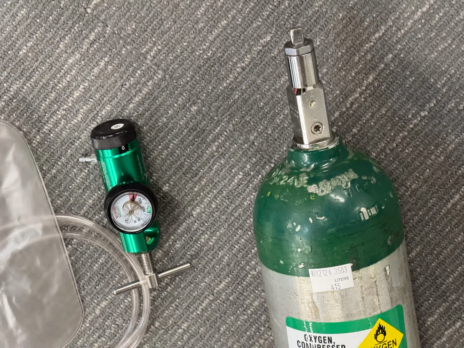 A green-topped portable medical oxygen cylinder labeled Oxygen, Compressed, lying beside a regulator with a pressure gauge and clear tubing