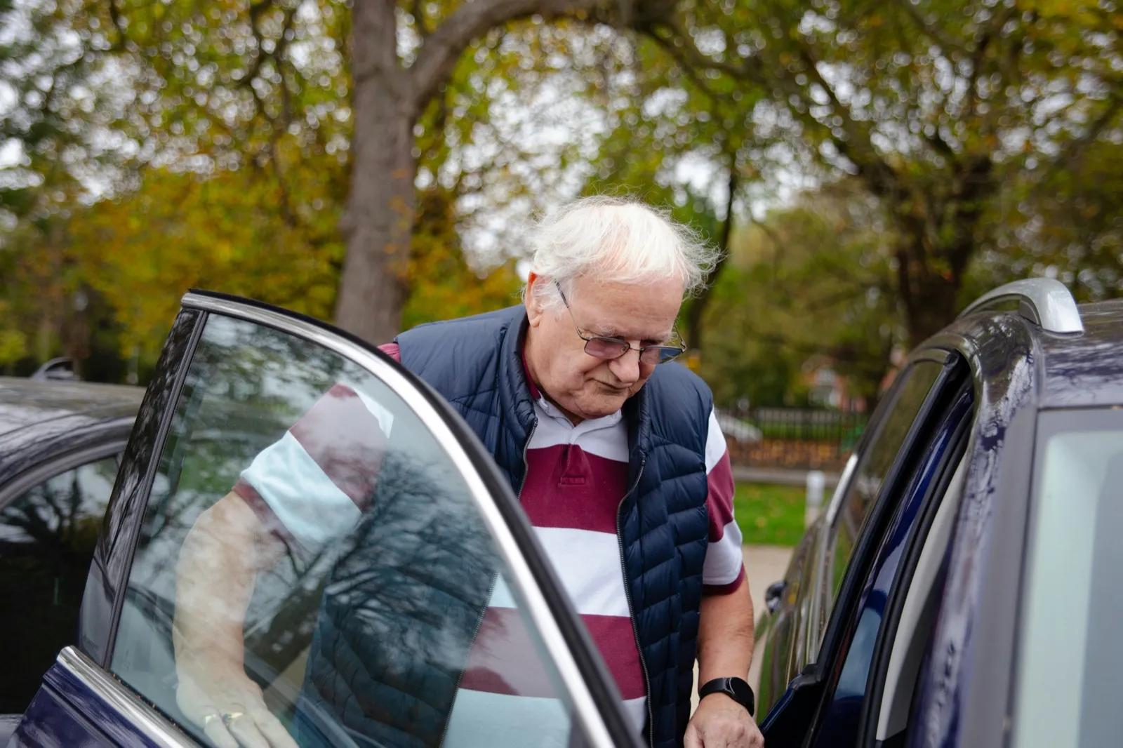 An older man with white hair and glasses leans in to get into a dark car through its open door, with trees behind him