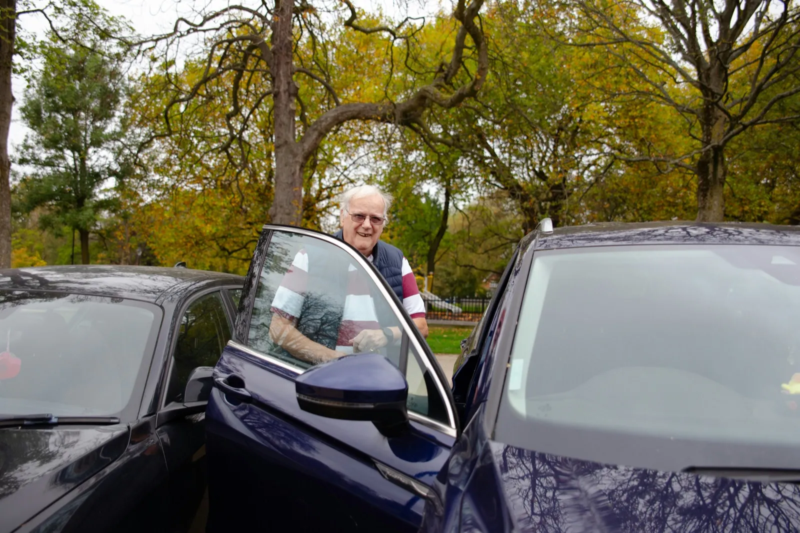 A smiling older man in glasses holds open the door of a dark blue car in a parking area, with autumn trees behind him
