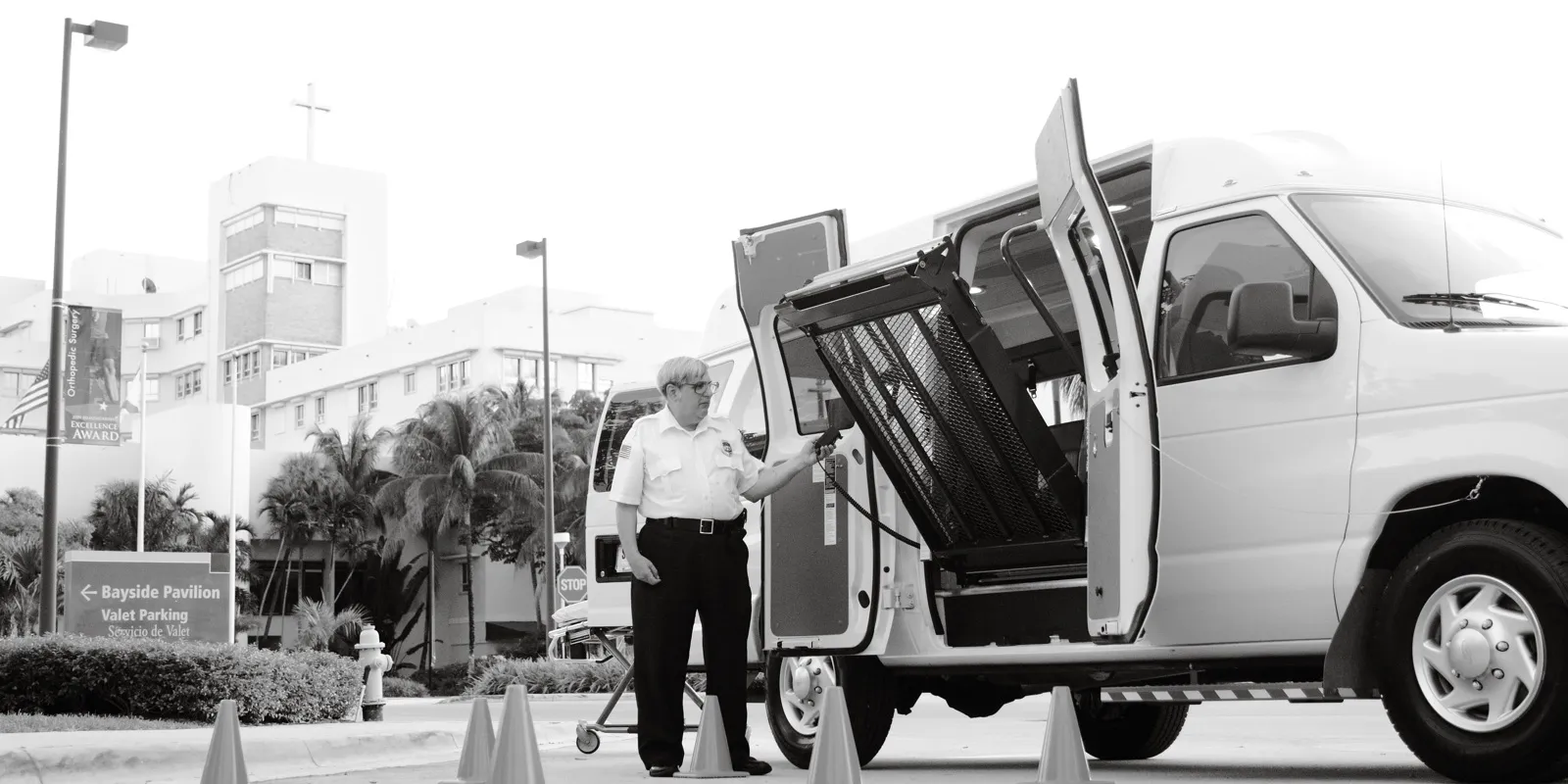 A driver in a uniform shirt uses the hand control to fold a wheelchair lift into the side door of a white high-roof van