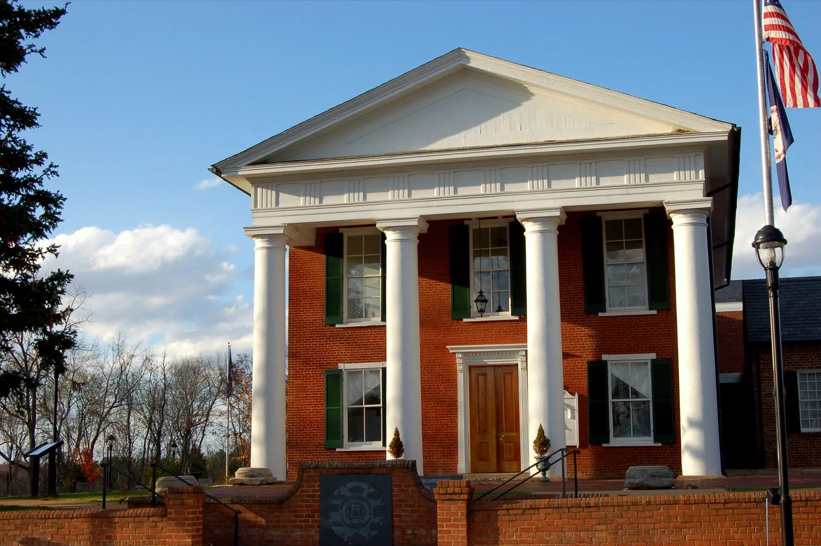 A red brick county courthouse in Virginia with four white columns, green shutters, and flags beside it under a blue sky