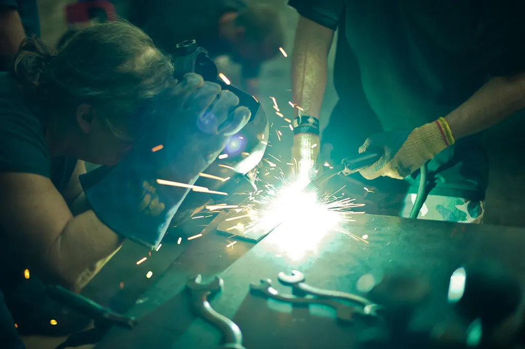 Two people in heavy gloves weld at a workbench in a welding class, with sparks flying and wrenches lying on the table