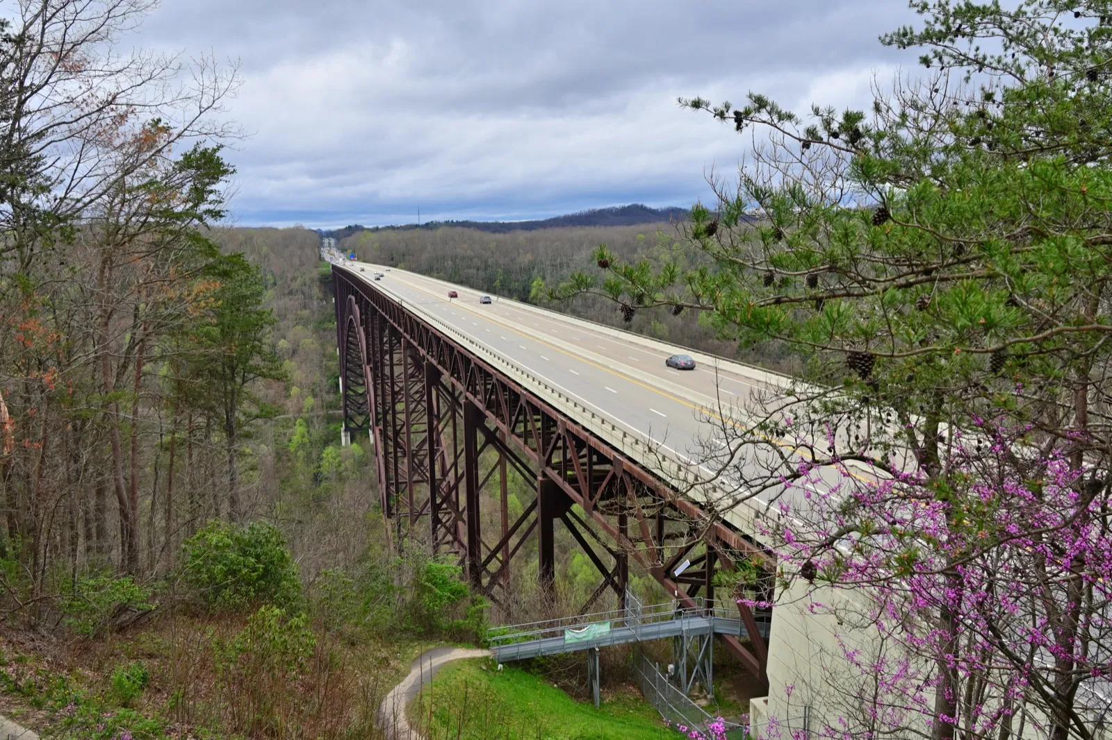 Cars crossing the New River Gorge Bridge on US 19, a long steel arch bridge over wooded hills in spring, seen from a trail beside it