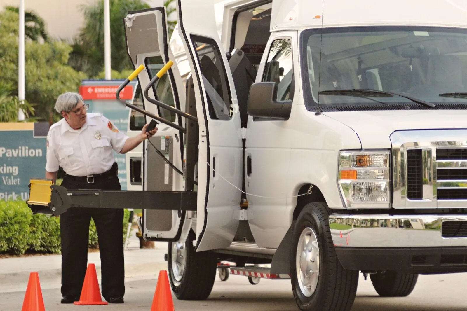A driver in a white uniform shirt works the controls of a wheelchair lift at the open side doors of a white high-roof van outside a hospital