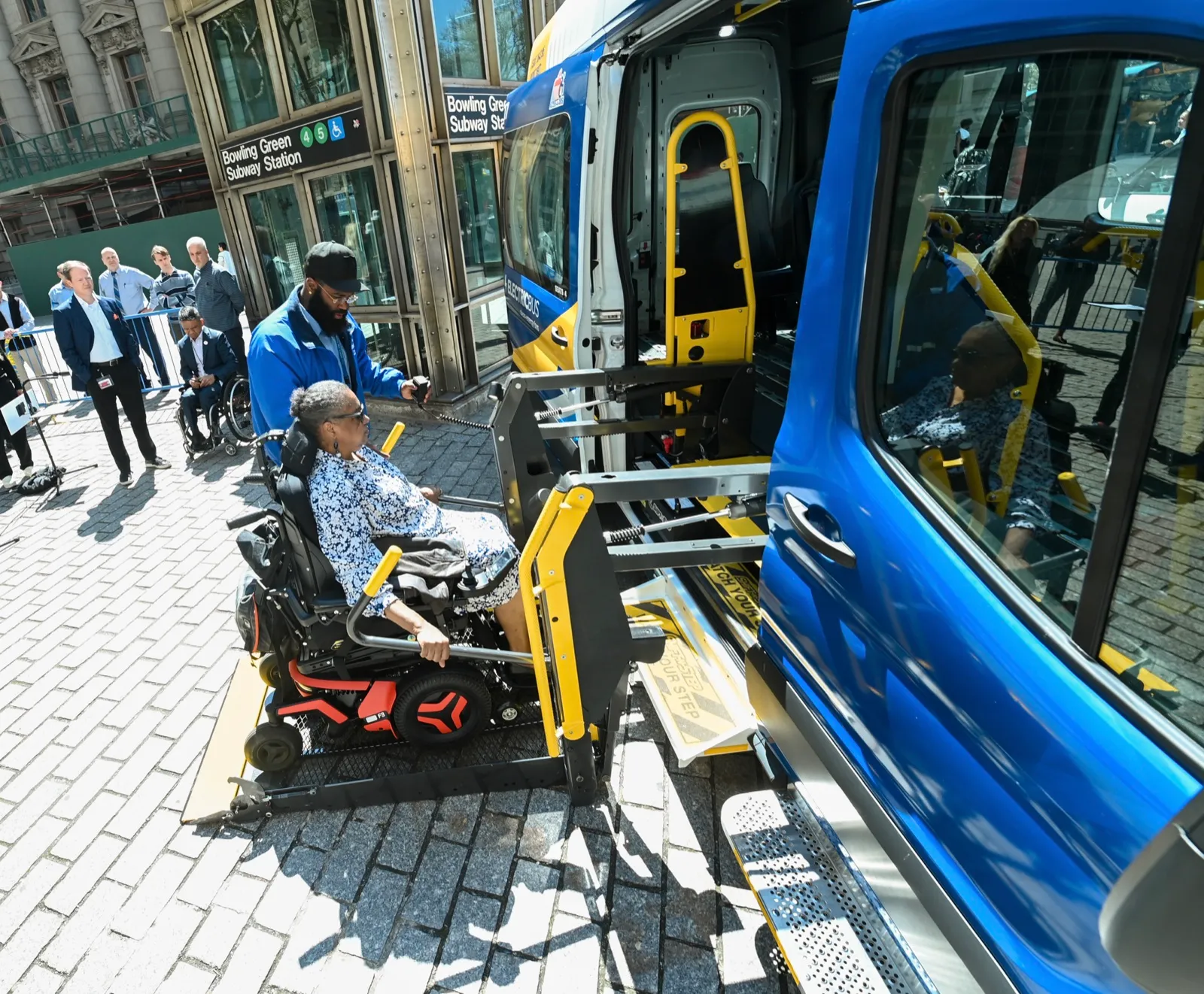 A woman in a power wheelchair rides a platform lift up to the side door of a full-size van while a man works the lift's hand control