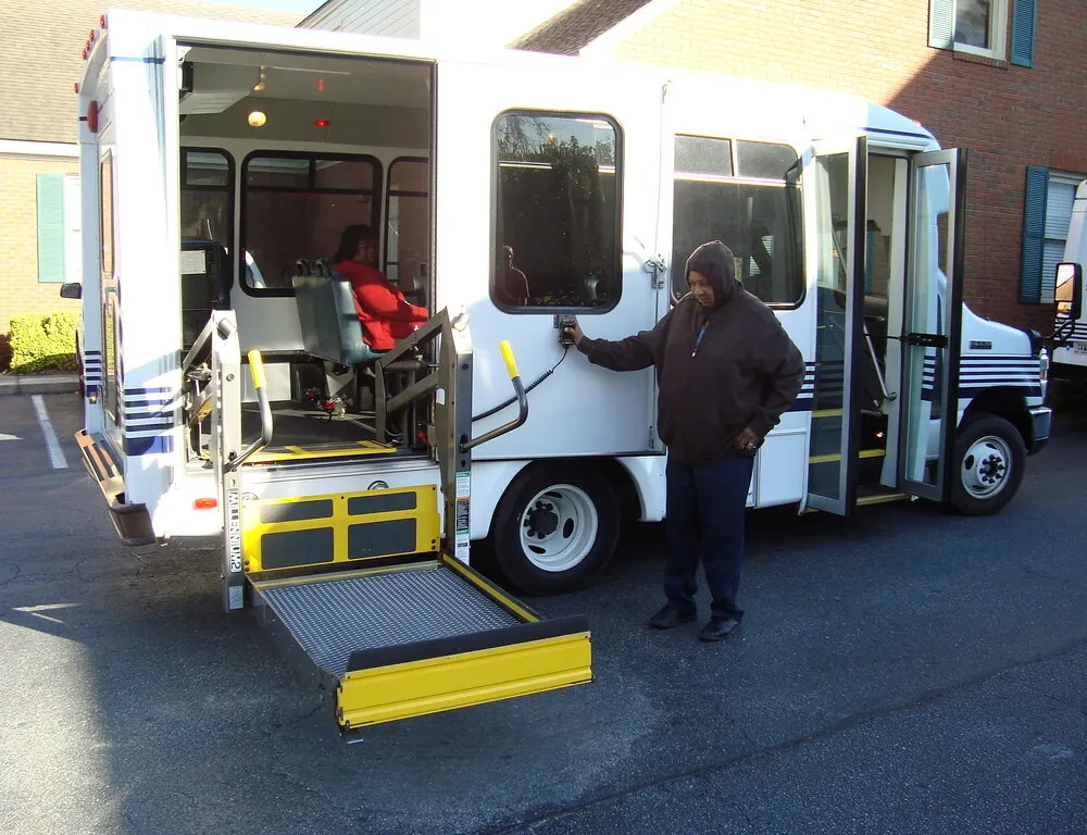 A person holds the hand control of a wheelchair lift lowered to the pavement at the side door of a white cutaway bus, with a rider seated inside