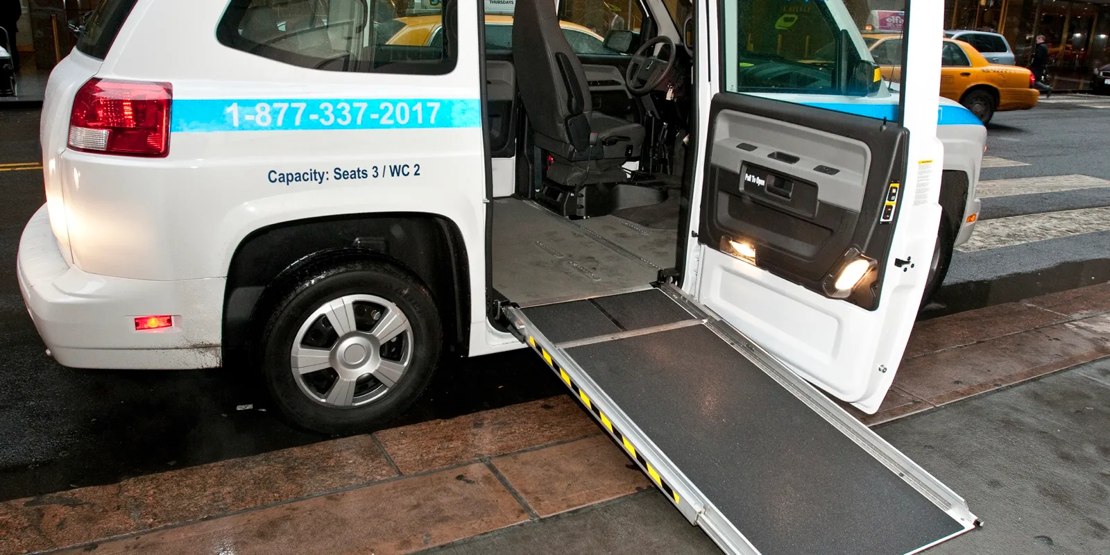 A white wheelchair accessible van at a city curb with its side door open and a fold-out ramp lowered to the sidewalk