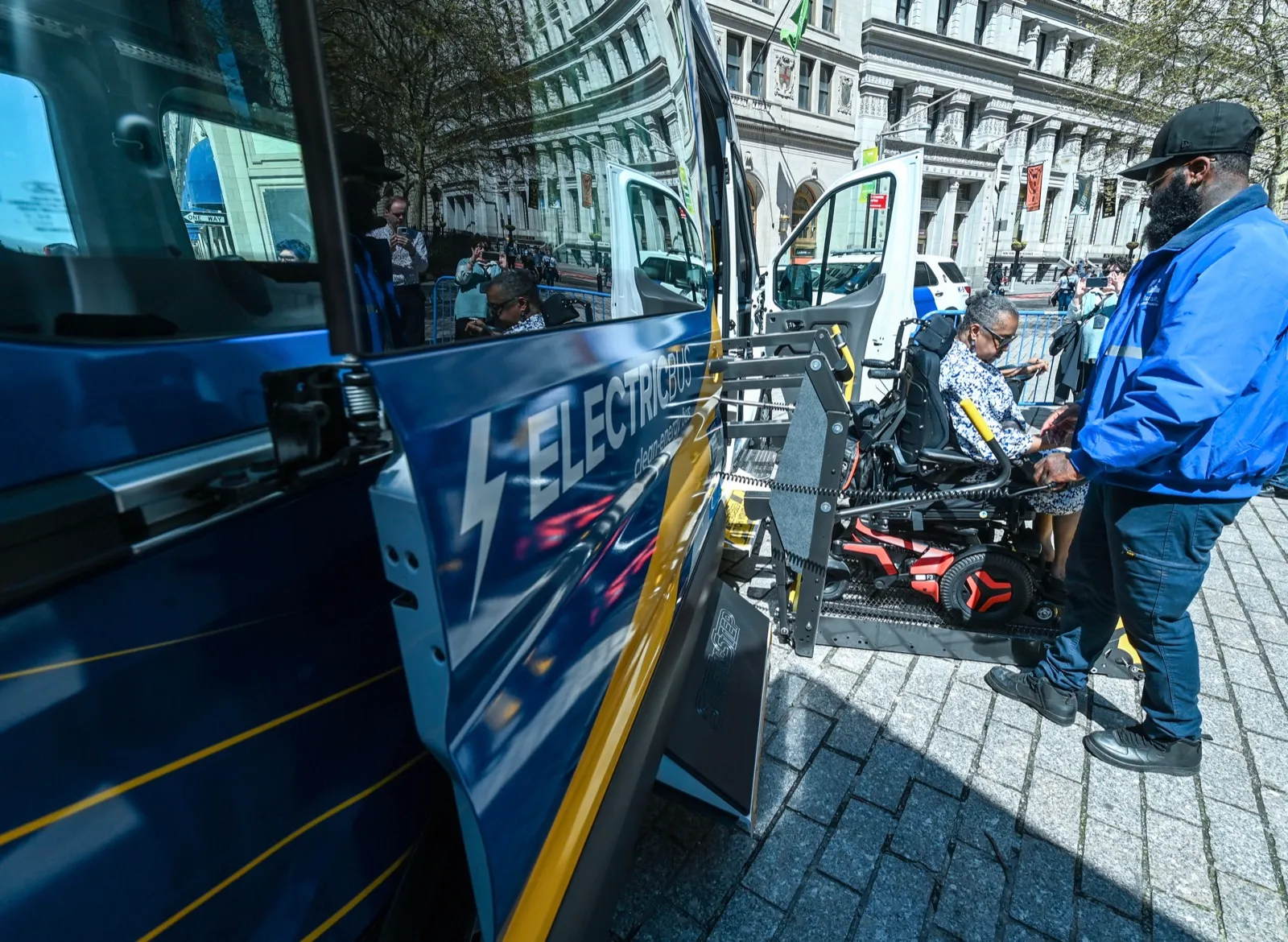 A man in a blue jacket runs the wheelchair lift of an accessible van while a woman in a power wheelchair rides the platform