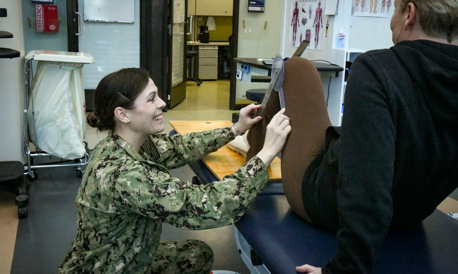 A smiling physical therapist in a Navy uniform kneels beside a patient on a treatment table and measures how far the knee bends