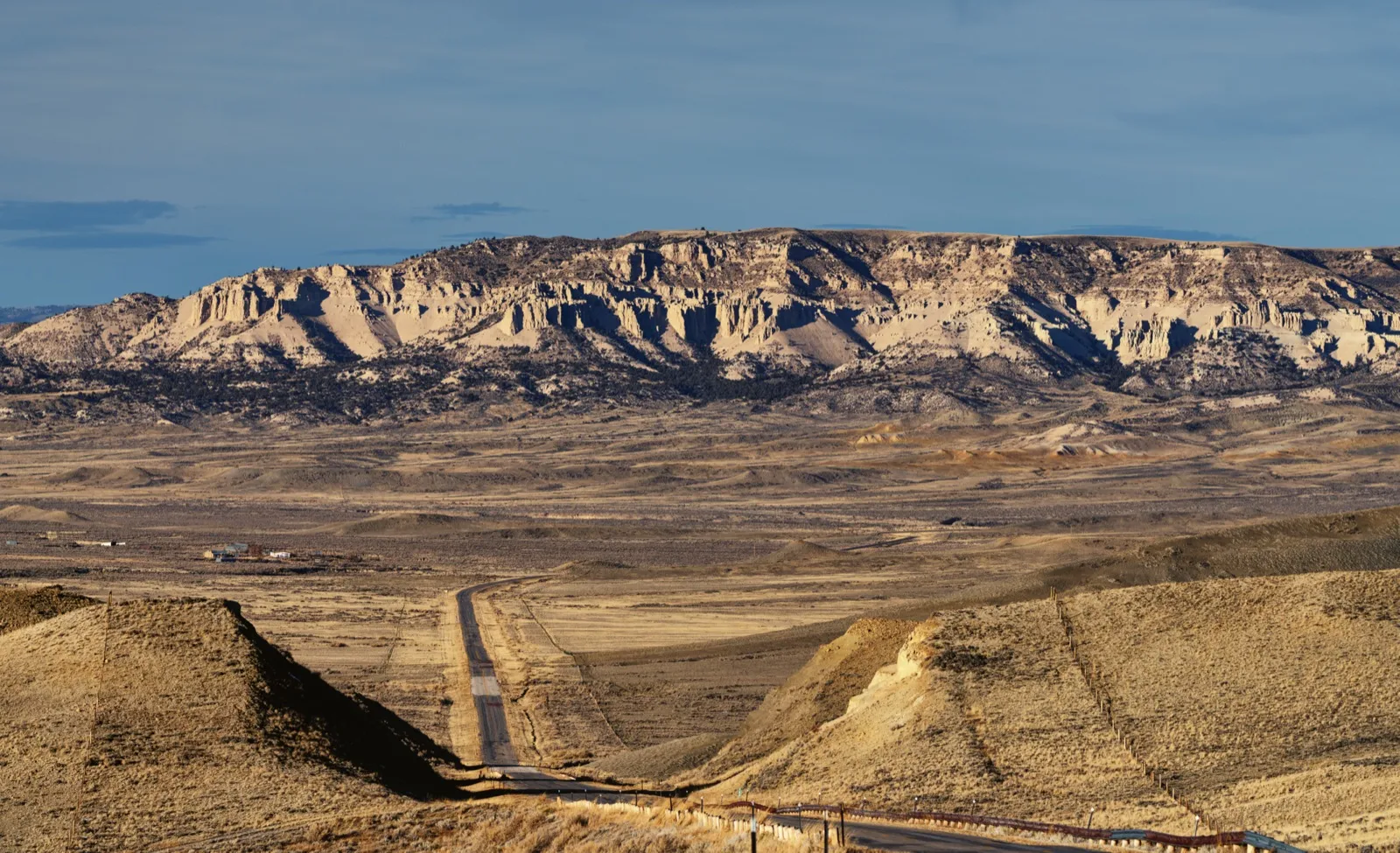 A two-lane highway running through dry hills toward pale cliffs in Carbon County, Wyoming, under a blue sky